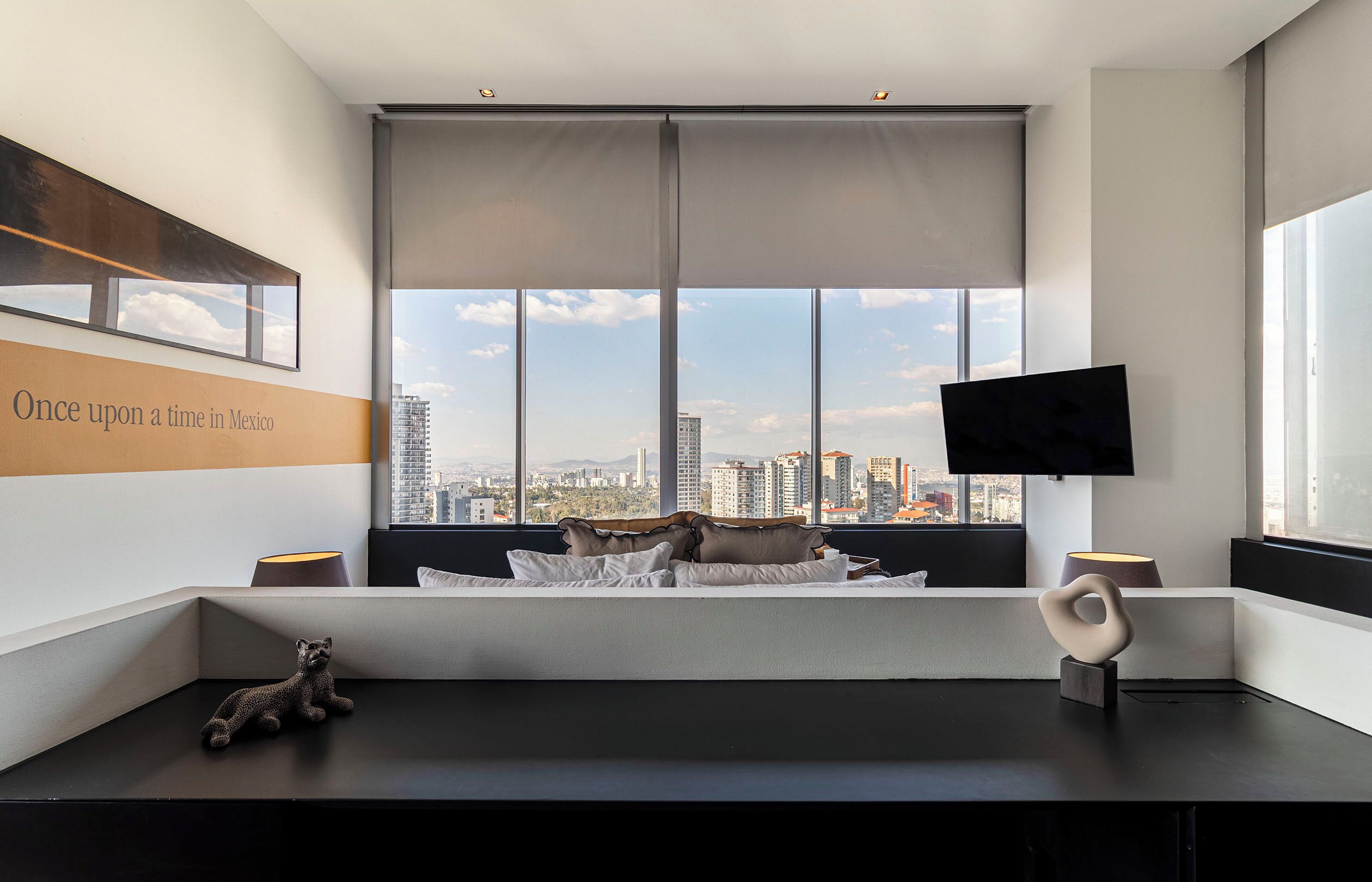 A modern hotel room with a large window offering a city view, a flat-screen TV mounted on the wall, and a cozy seating area with pillows and a decorative sculpture.