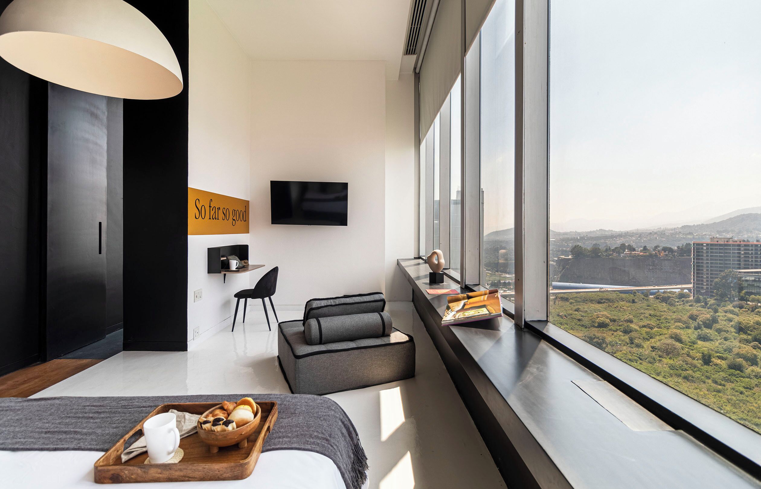 A modern hotel room with a large window, a flat-screen TV, a sofa, a dining table with breakfast, and a city view.