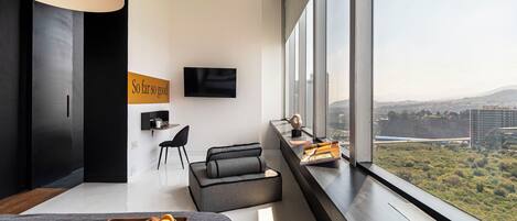 A modern hotel room with a large window, a flat-screen TV, a sofa, a dining table with breakfast, and a city view.