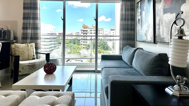Un salon moderne avec un canapé gris, une table basse blanche et une vue sur la ville grâce à de grandes fenêtres.