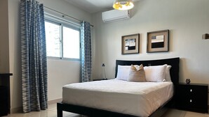 Un lit bien fait, avec une tête de lit noire, une table de chevet avec une lampe et une fenêtre avec des rideaux.