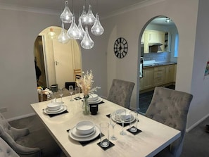 A dining area with a marble table set for four, surrounded by tufted chairs. A modern chandelier hangs above, and a wall clock is visible. - Beautiful 3-bed Apartment in Swansea (Swansea)