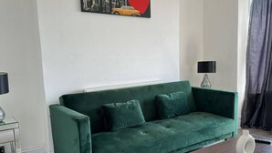 A green sofa with two cushions, a small table with a lamp, and a painting on the wall. - Newport City Escape - 2 Bedroom Apartment (Newport)