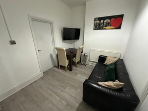 A small living room with a black leather sofa, a dining table with chairs, a flat-screen TV mounted on the wall, and a radiator.