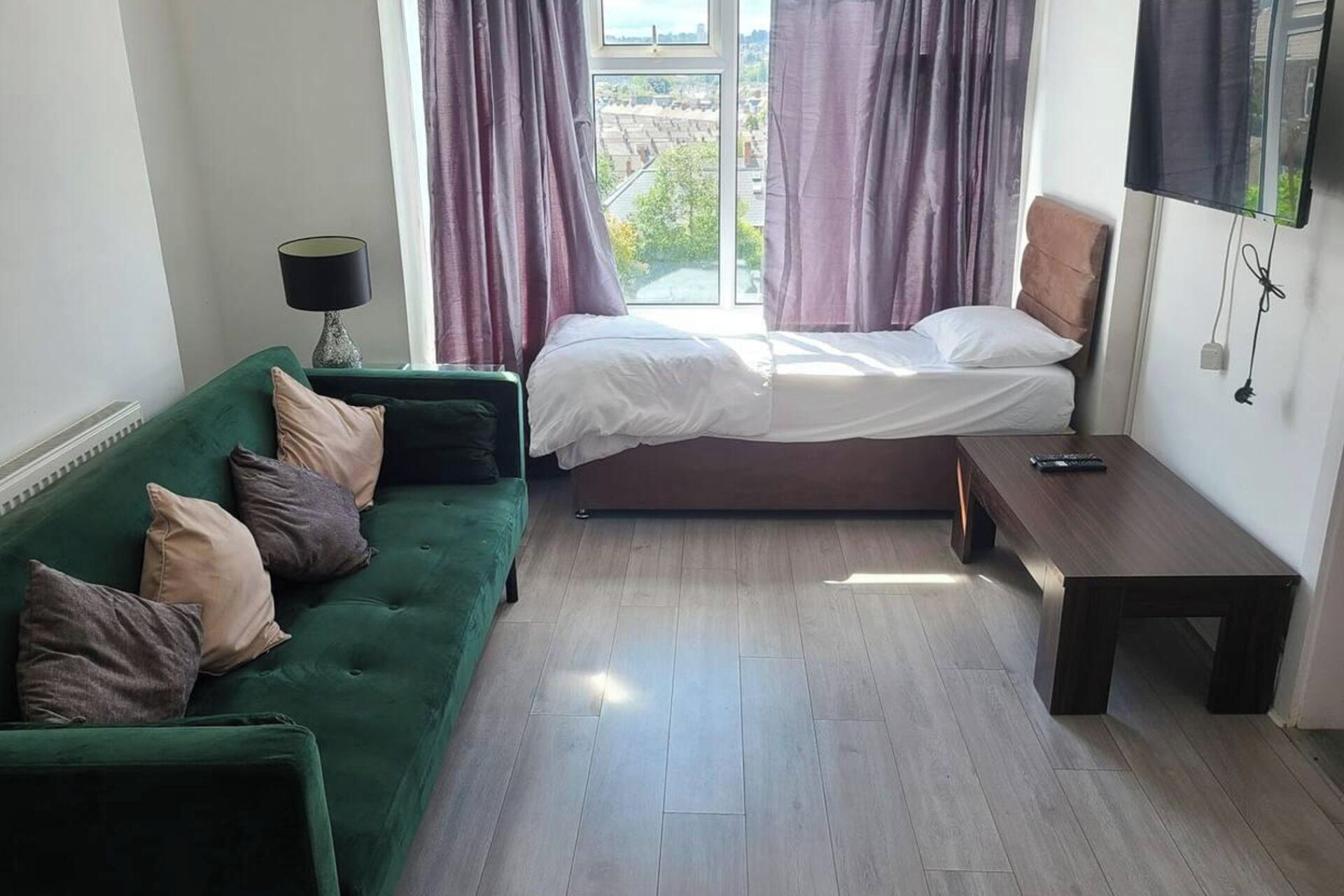 Una stanza piccola e luminosa con un divano verde, un letto singolo, un comodino in legno e una finestra con tende.