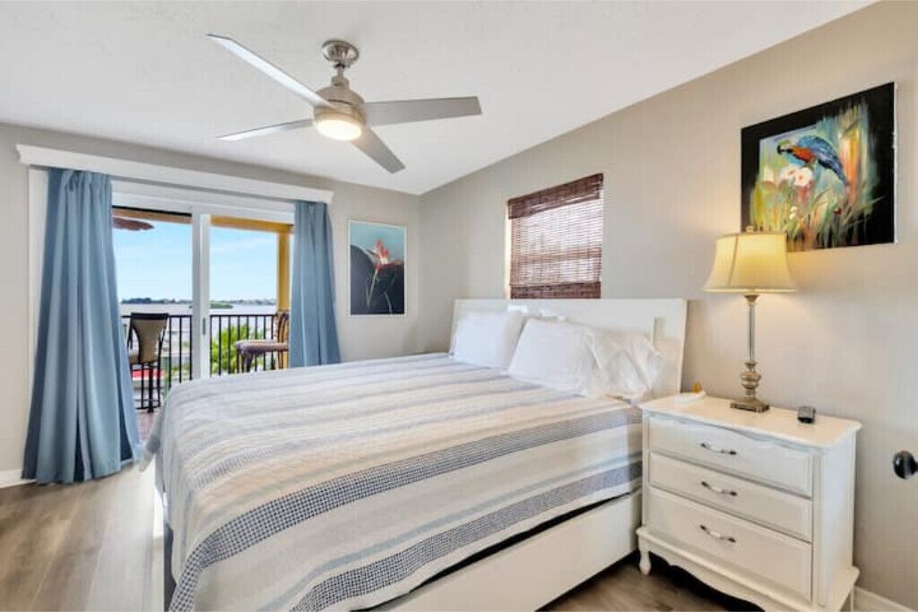 Une chambre à coucher avec un lit, une table de chevet, un ventilateur de plafond, une fenêtre avec des rideaux et un tableau au mur.
