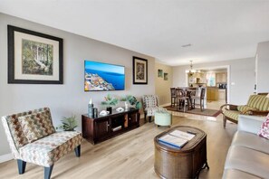 Sala de estar con televisor de pantalla plana, comedor con mesa y sillas, y cocina al fondo.