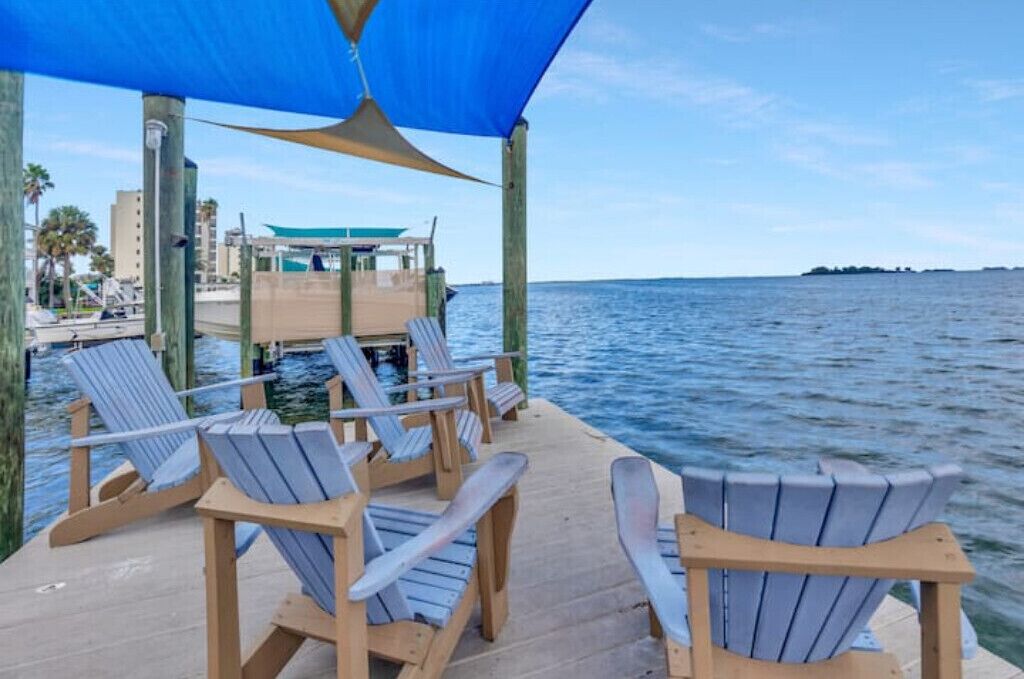 Een houten terras met Adirondack-stoelen met uitzicht op kalm water, een overdekt gebied met een blauw zonnescherm en een verre weergave van een gebouw en de kustlijn.