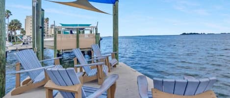 A wooden deck with Adirondack chairs overlooking calm water, a covered area with a blue canopy, and a distant view of a building and shoreline.
