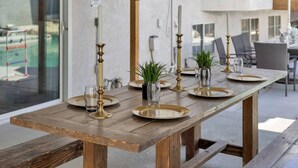 Ein Holztisch mit goldenen Tellern, Gläsern und Kerzenständern, platziert in einem modernen Essbereich im Freien mit Blick auf einen Pool und Gebäude.