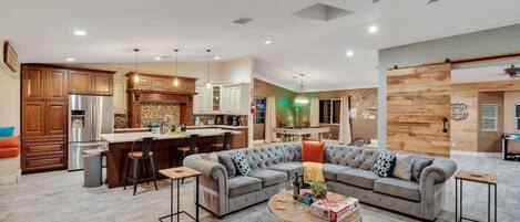 A modern living room with a sectional sofa, a coffee table, and a kitchen area with wooden cabinets and a stainless steel refrigerator.