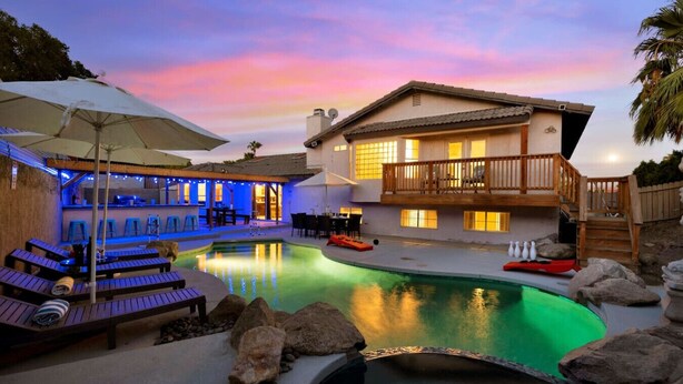 Poolbereich mit Liegestühlen und Sonnenschirmen, ein Haus mit Balkon und ein Sonnenuntergangshimmel.