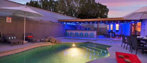 A swimming pool with lounge chairs and a bar area, surrounded by umbrellas and string lights.
