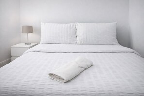 A neatly made bed with white bedding and a rolled towel, a bedside lamp, and a white nightstand. - 4-bed House in Enfield- Sleeps 8- Private Garden (Enfield)