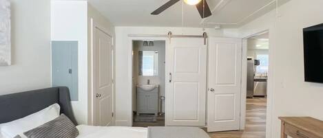 A bedroom with a bed, a ceiling fan, a TV, and a view into the kitchen.