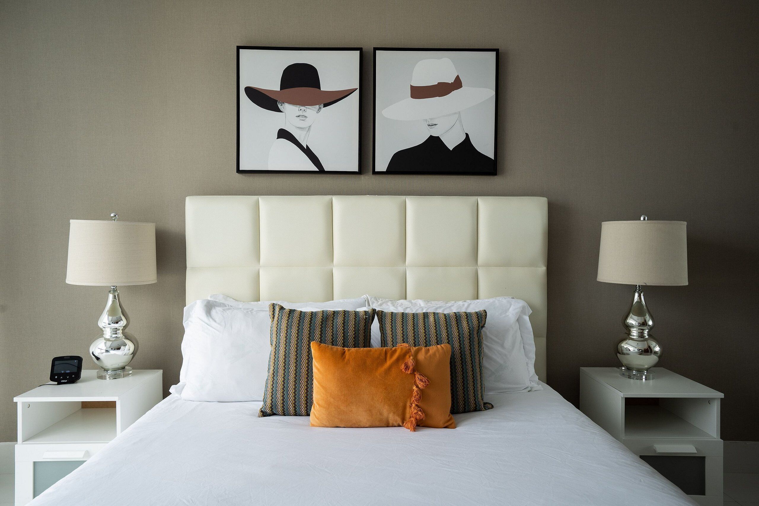 Una cama bien hecha con almohadas blancas y estampadas, dos mesitas de noche con lámparas y dos obras de arte enmarcadas en la pared.