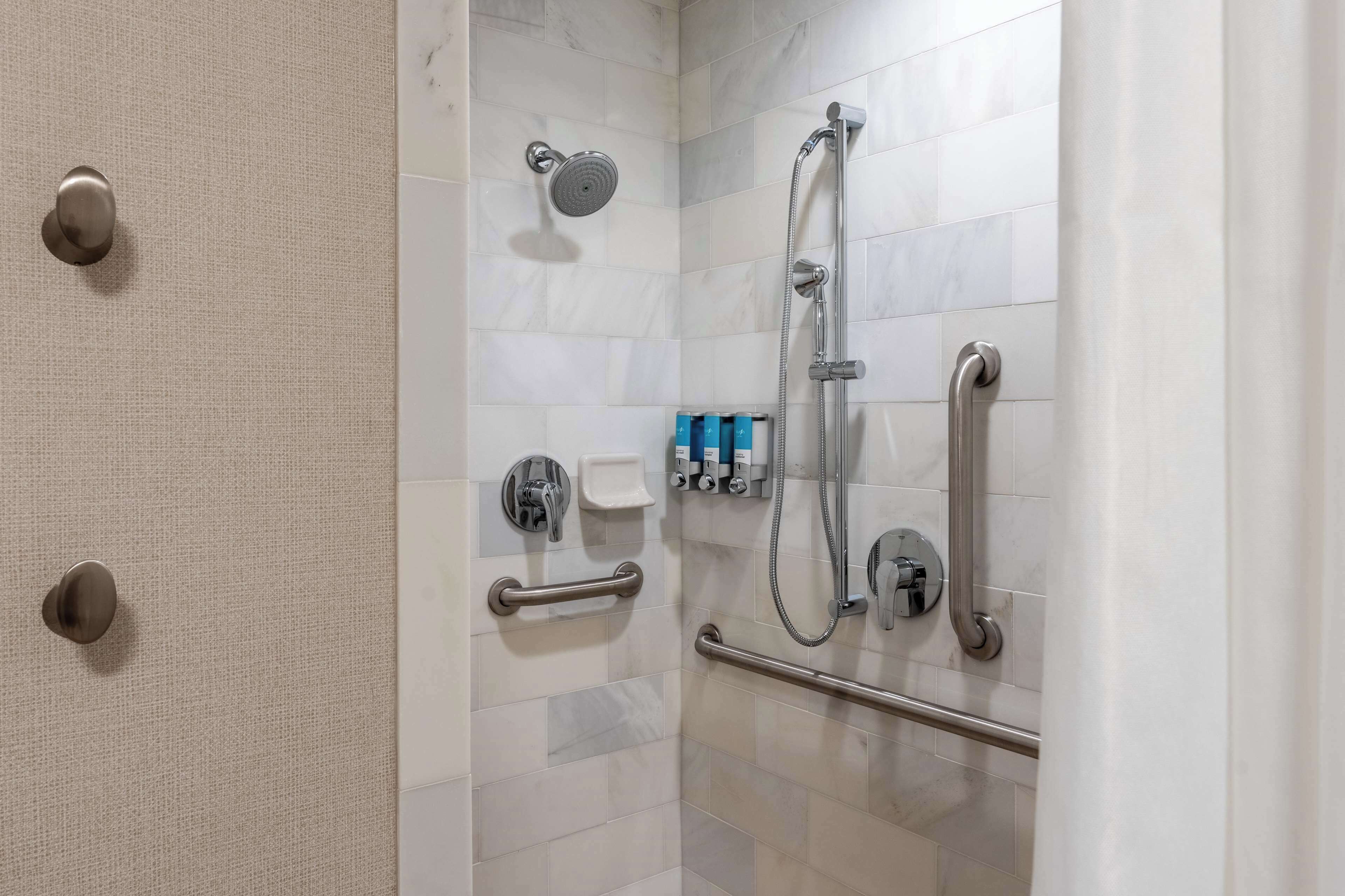 Een douchegedeelte met een handdouche, een douchekop aan de muur en een flexibele slang. Er zijn handgrepen en een veiligheidsbeugel voor extra zekerheid.