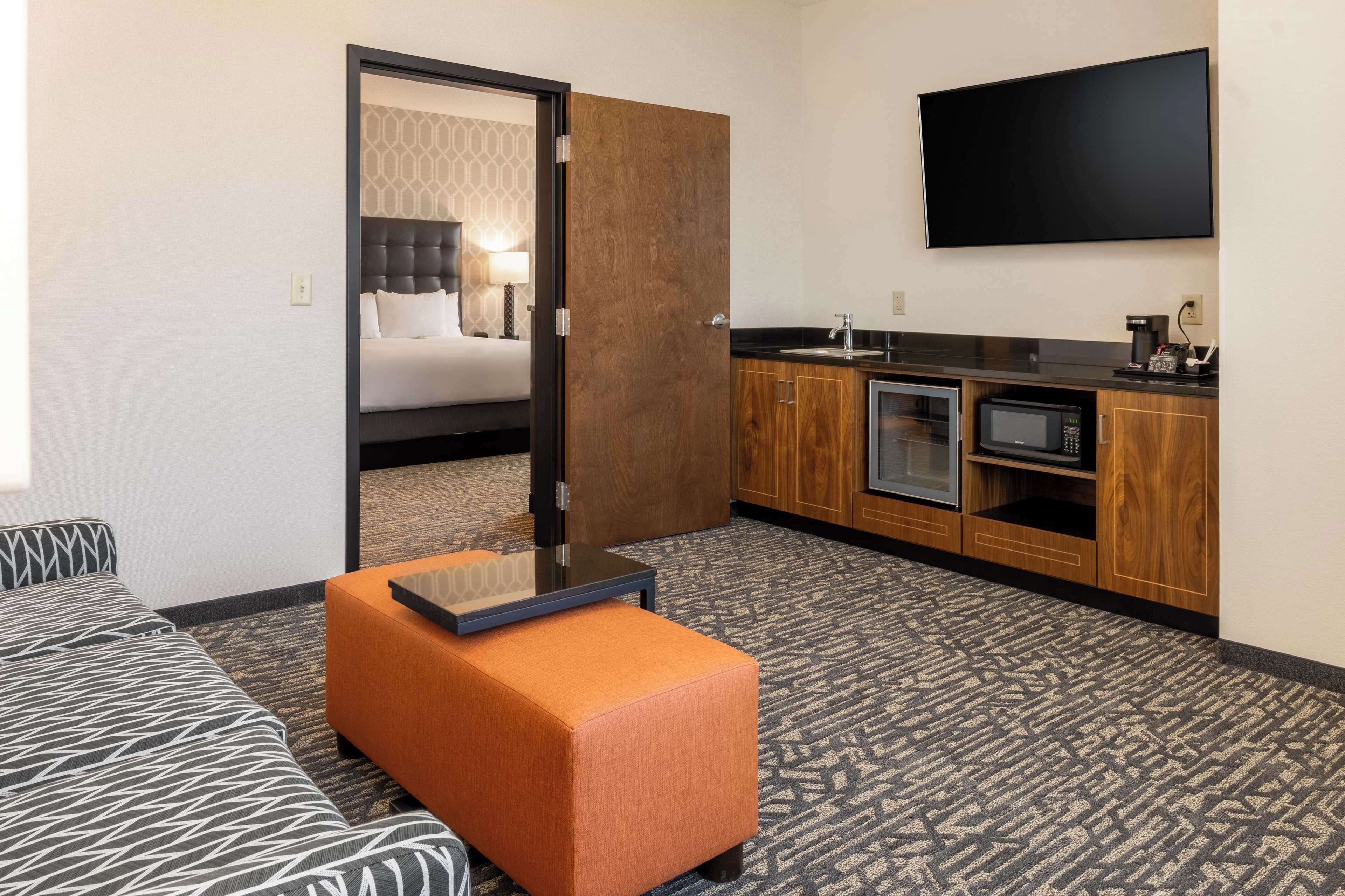 Habitación de hotel con cama, sofá, reposapiés naranja, kitchenette con microondas y televisor montado en la pared.