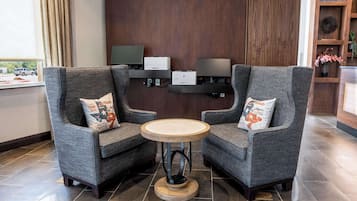 Two grey armchairs with matching cushions, a round wooden table, and a dark wood paneling wall with two monitors and printers.