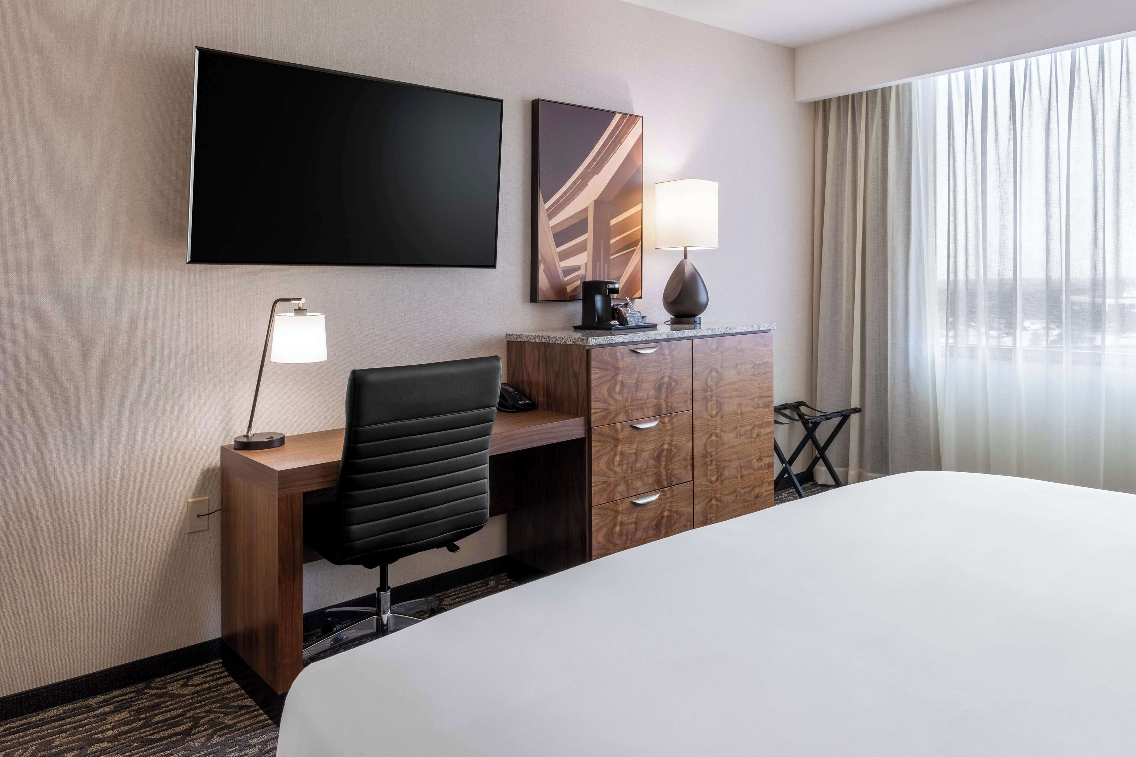 Een hotelkamer met een bed, een bureau met stoel, een televisie, een lamp, een koffiezetapparaat en uitzicht op de stad.