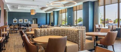 Een moderne eetruimte met ronde tafels en zitbanken, grote ramen en een plafond met ronde lampen.
