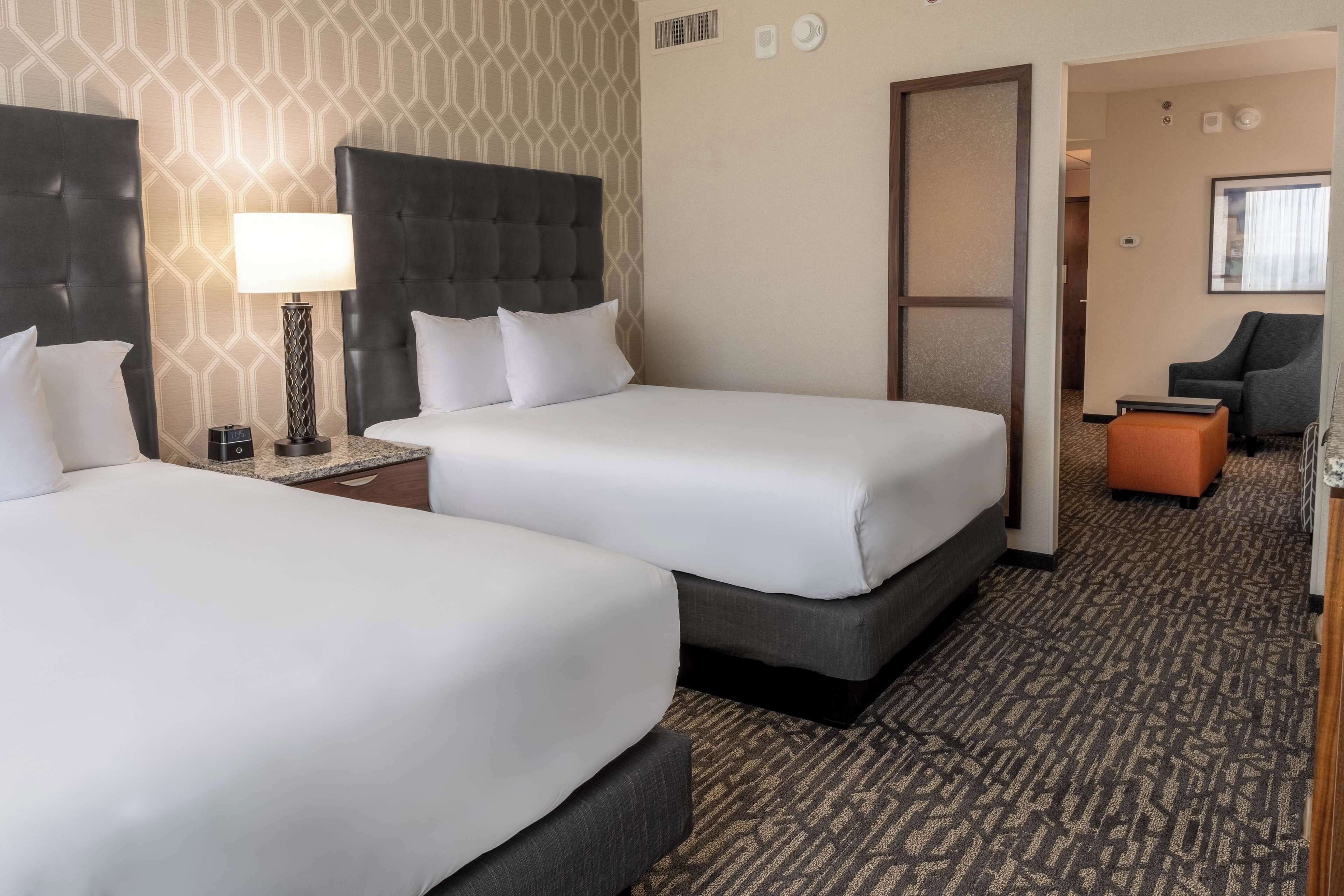 A hotel room with two beds, a nightstand, a lamp, and a seating area with a chair and ottoman.