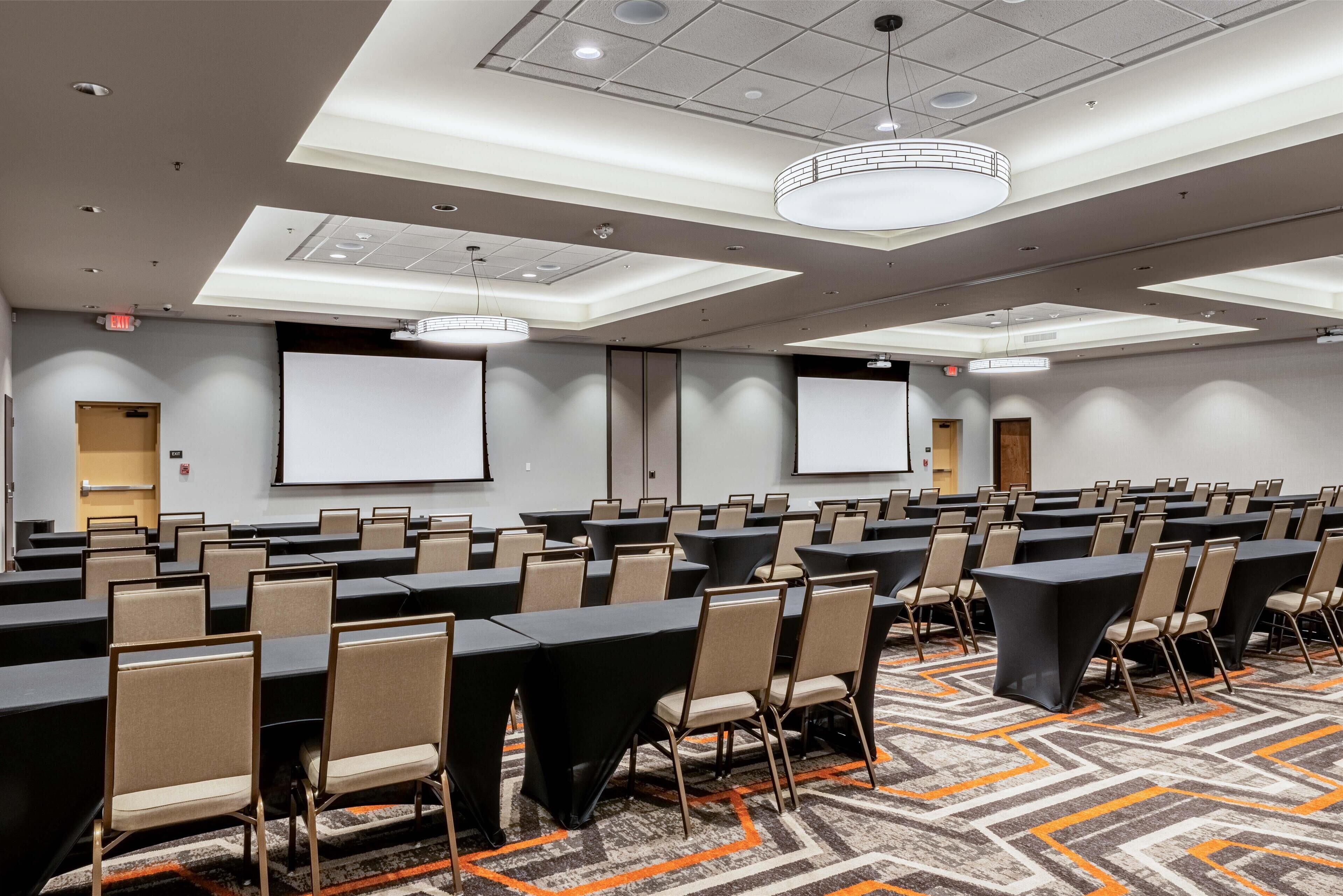 A conference room with multiple tables and chairs, a large screen, and a patterned carpet.