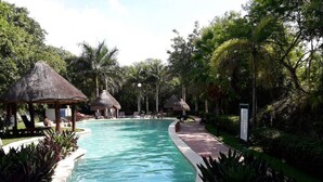 A swimming pool surrounded by tropical vegetation and thatched-roof structures. - Cozy Apartment 1B Puerto Morelos  (PUERTO MORELOS)