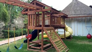 A wooden play structure with a slide and swings, set against a backdrop of a thatched-roof hut and lush greenery. - Cozy Apartment 1B Puerto Morelos  (PUERTO MORELOS)