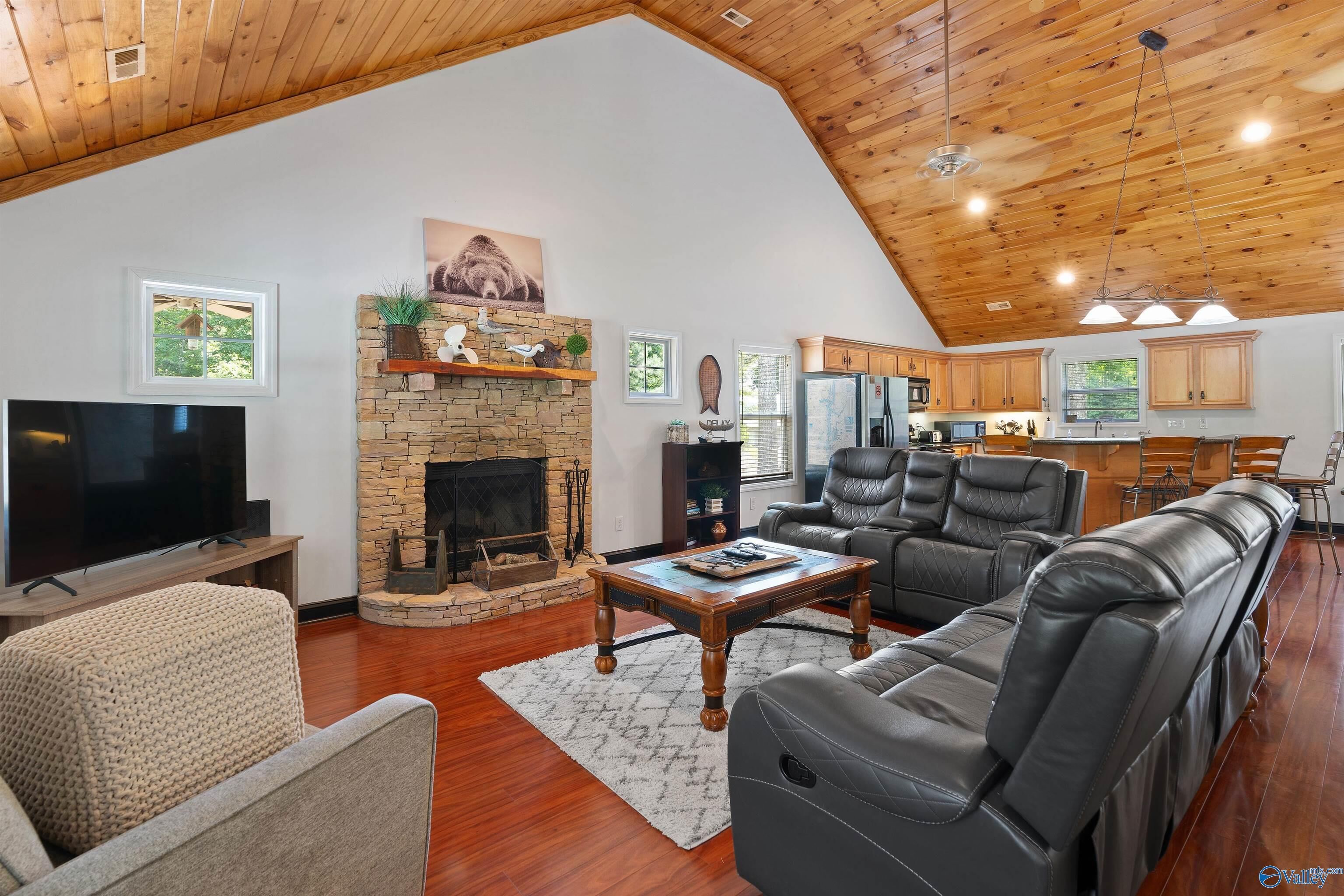 A spacious living room with a stone fireplace, a large flat-screen TV, leather sofas, and a kitchen area with wooden cabinets.