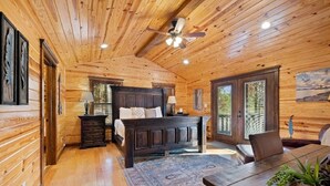Ein Holzblockhaus-Interieur mit Holzbalkendecke, einem Bett mit dunklem Kopfteil, einem Esstisch und einem großen Fenster mit Blick auf Bäume.