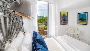 Een slaapkamer met een bed, een witte stoel en een balkon met uitzicht op groen en een gebouw.