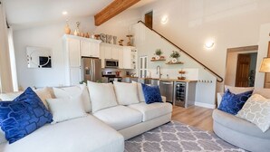 Un salon moderne avec un canapé blanc, un tapis à motifs gris et un escalier menant à un espace cuisine doté de meubles blancs et d’appareils électroménagers en acier inoxydable.