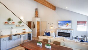 Une cuisine moderne avec un plafond en bois, un coin repas avec une table en bois, un salon avec un canapé et une cheminée, et une grande fenêtre donnant sur un paysage enneigé.