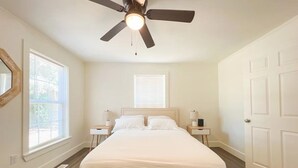 Un dormitorio con una cama, un ventilador de techo, dos mesitas de noche con lámparas, una ventana y una puerta.