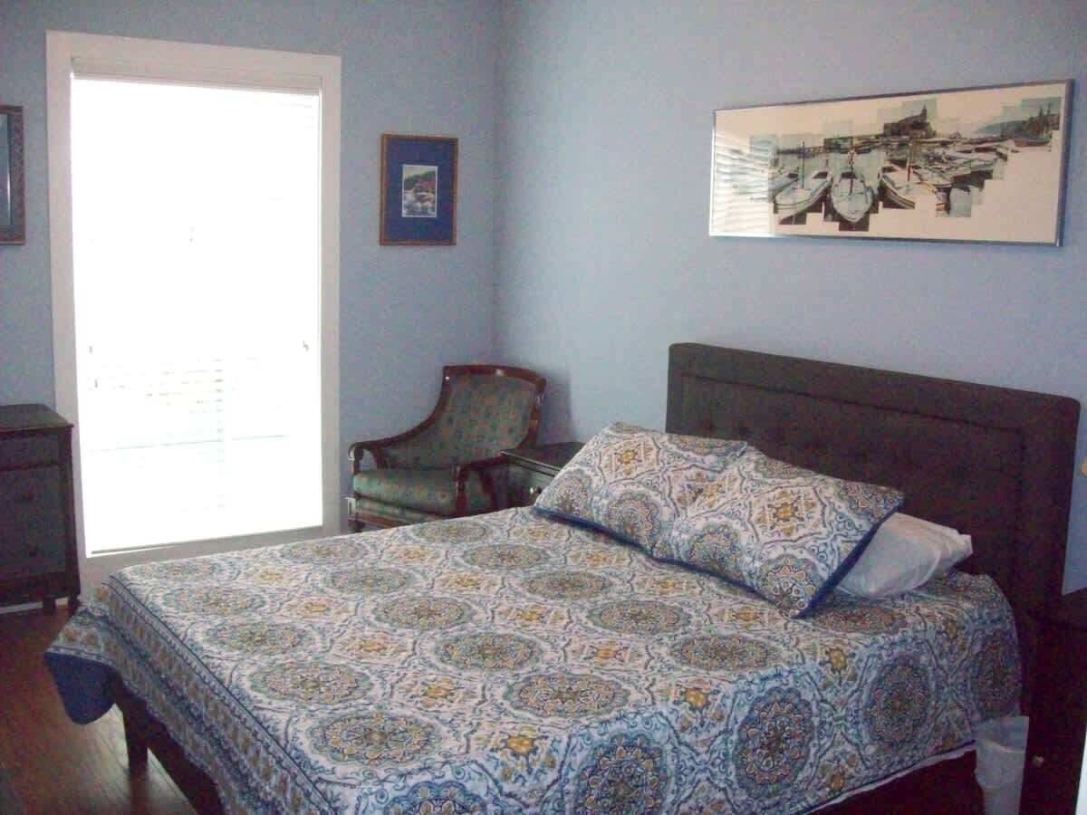 A bedroom with a patterned bedspread, a chair, a framed picture, and a window with blinds.