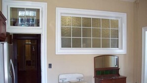 Een kamer met een houten vloer, een witte deur, een raam met geëtste glassering, een houten dressoir en een spiegel.