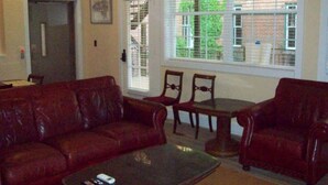A living room with a dark brown leather sofa, a coffee table, and two wooden chairs. A window with blinds is visible, and a door is in the background. - Large Historic Downtown Loft - Rear (Macon)