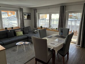 Un salon moderne avec un canapé gris, une table à manger avec des chaises et de grandes fenêtres avec des rideaux.