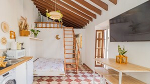 Quarto compacto e bem organizado, com uma cama, pequeno espaço de cozinha, escada de madeira, mesa e uma janela grande.