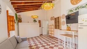 Uma sala de estar com piso xadrez, um sofá, uma TV e uma área de cozinha com geladeira e prateleiras de madeira.