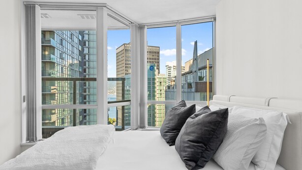 Une chambre moderne avec une grande fenêtre, du linge de lit blanc et une vue sur les bâtiments de la ville.