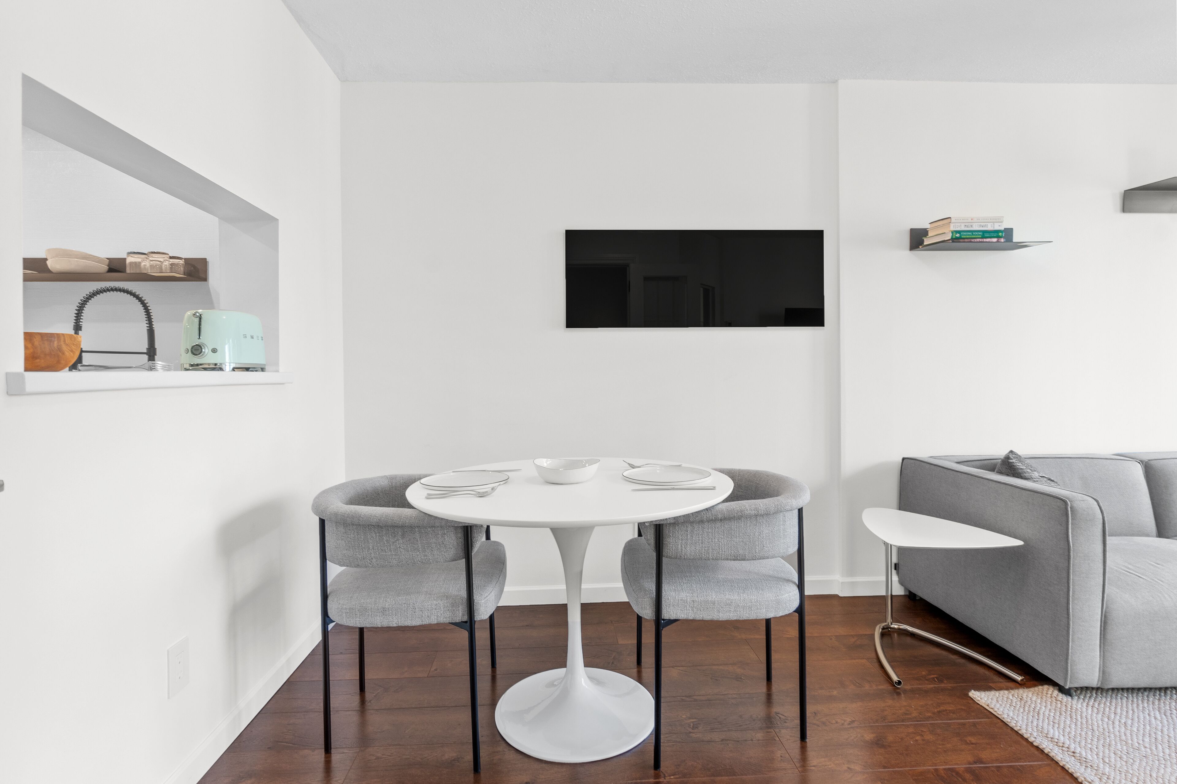 A modern living room with a round white table, grey armchairs, a grey sofa, a flat-screen TV mounted on the wall, and a small shelf with books and a kettle.