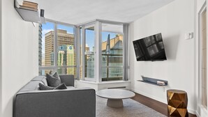 Un salon moderne avec un canapé gris, une table basse ronde, un téléviseur fixé au mur et une grande fenêtre donnant sur la ville.