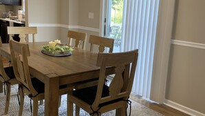 A dining area with a wooden table and chairs, a chandelier, a kitchen in the background, and a view through a window with blinds.