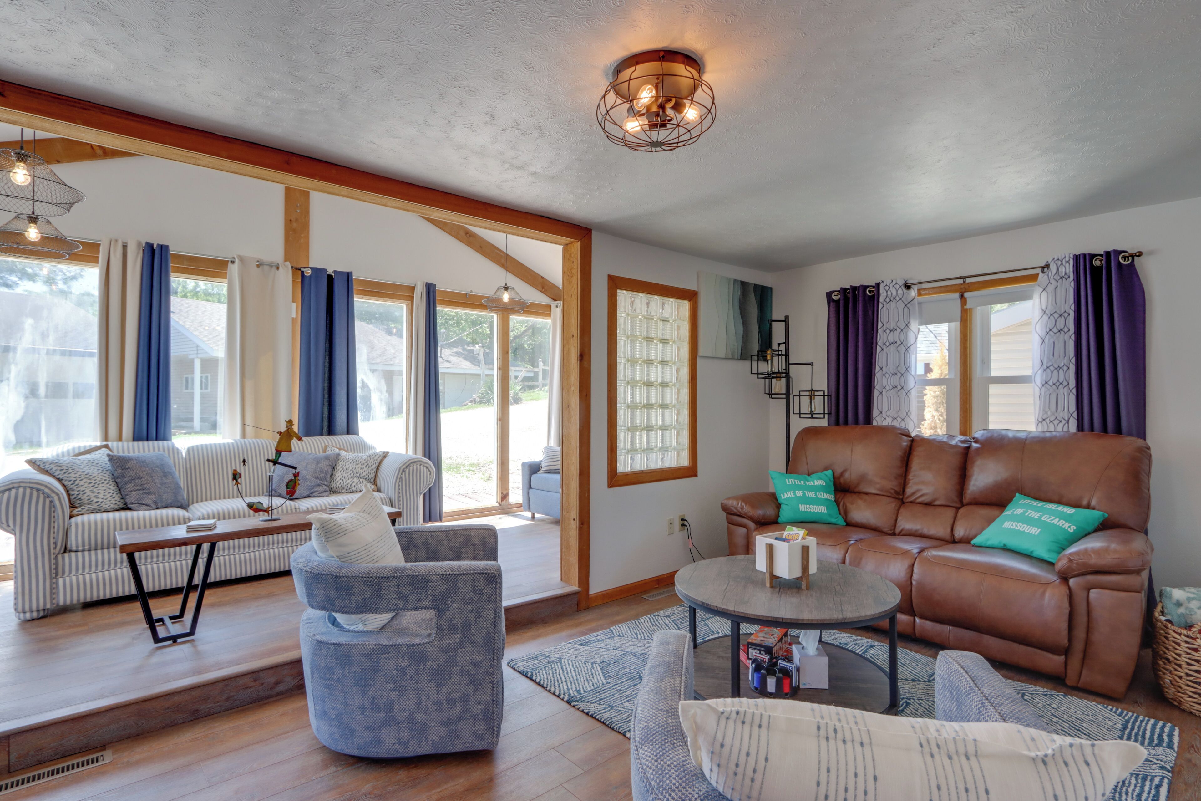 A spacious living room with a sofa, armchair, and coffee table, large windows with curtains, and a wooden ceiling.
