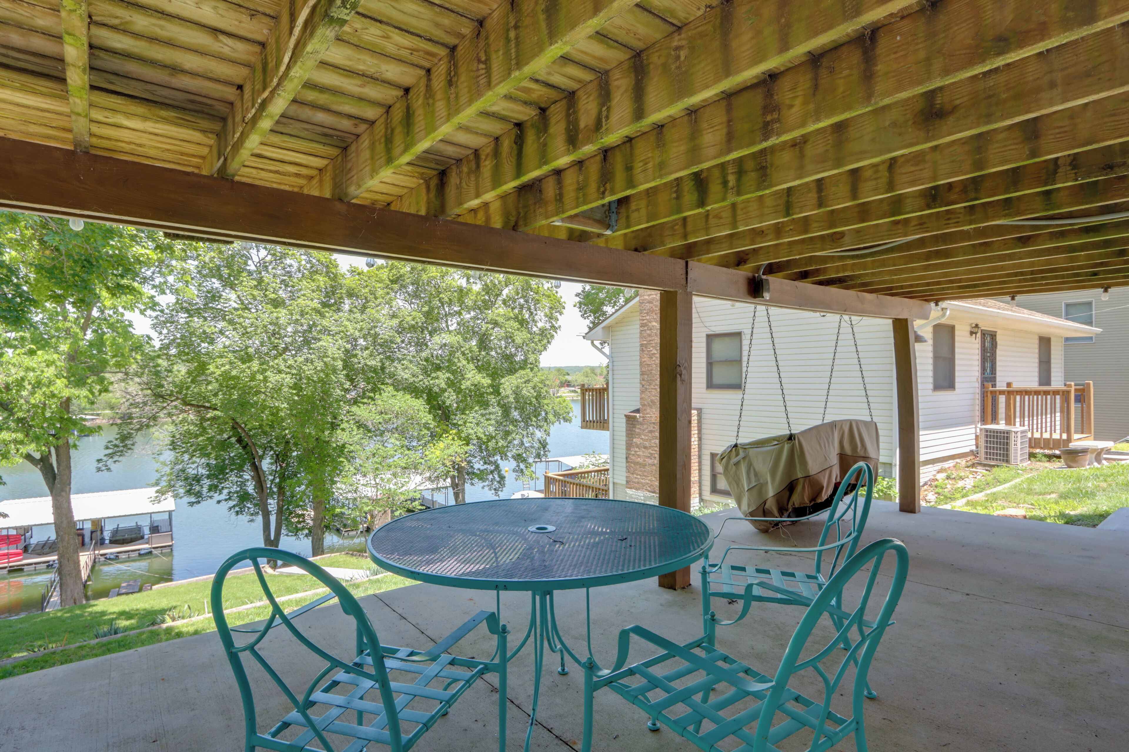 Een overdekt terras met een ronde tafel en stoelen, een schommel en uitzicht op een huis en bomen.