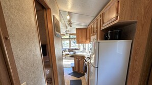 A compact RV kitchen with wooden cabinets, a stainless steel refrigerator, and a black mat on the floor. - Cozy Park Model minutes from many of UPs popular attractions & UTV/ATV Snow Trls (White Pine)