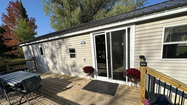 A single-story house with a deck, a sliding glass door, and a small porch area with a table and chairs. - Cozy Park Model minutes from many of UPs popular attractions & UTV/ATV Snow Trls (White Pine)