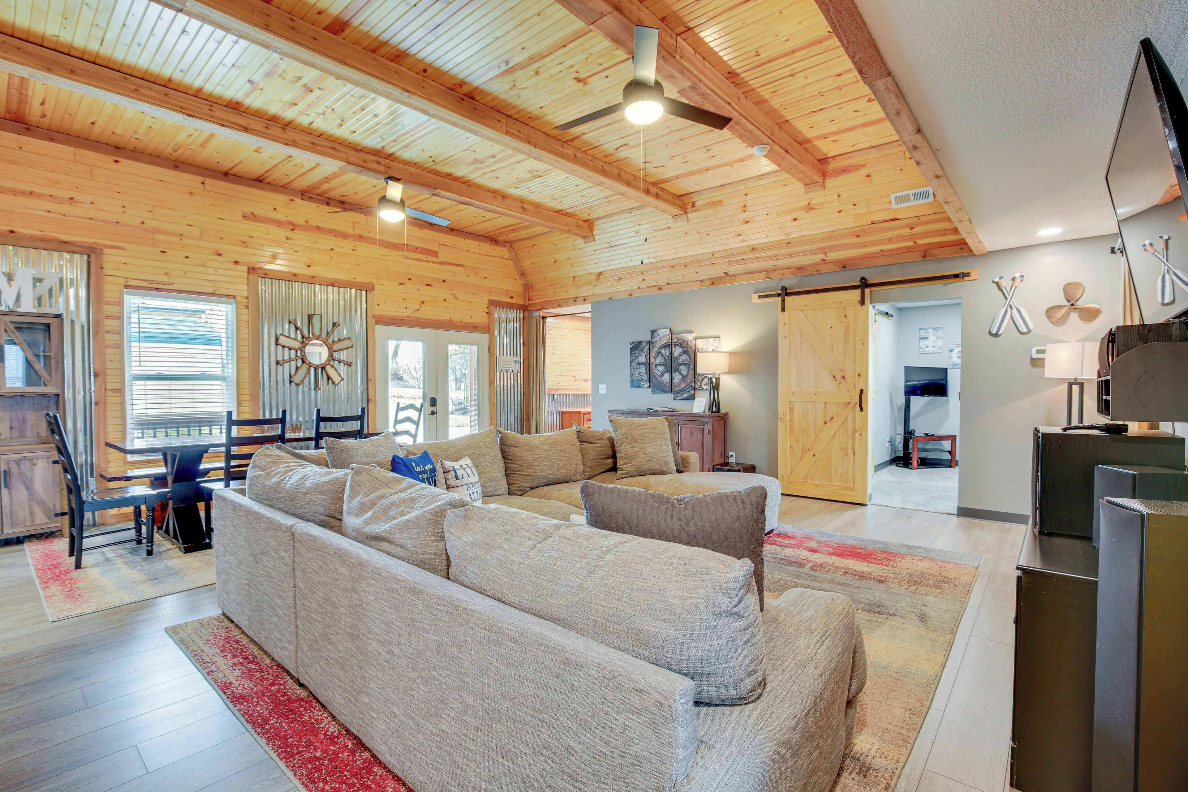 A spacious living room with a large beige sectional sofa, wooden ceiling beams, and a ceiling fan.